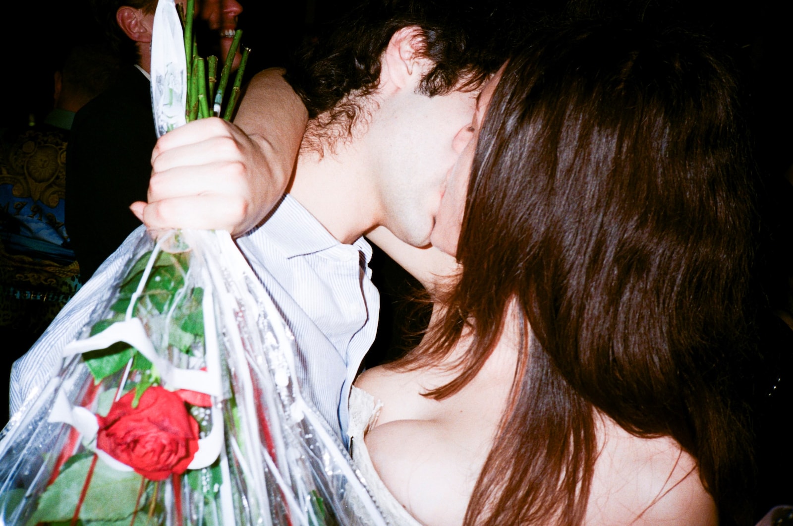 A film photograph of two people in close contact, one holding a translucent cellophane-wrapped bouquet with red roses and green stems against a dark background.