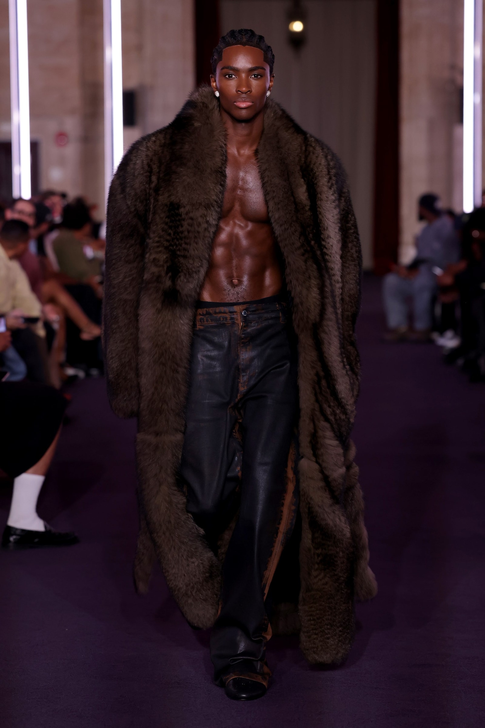 A Black male model walks the VIVET runway bare-chested beneath an oversized brown fur coat with dark leather trousers