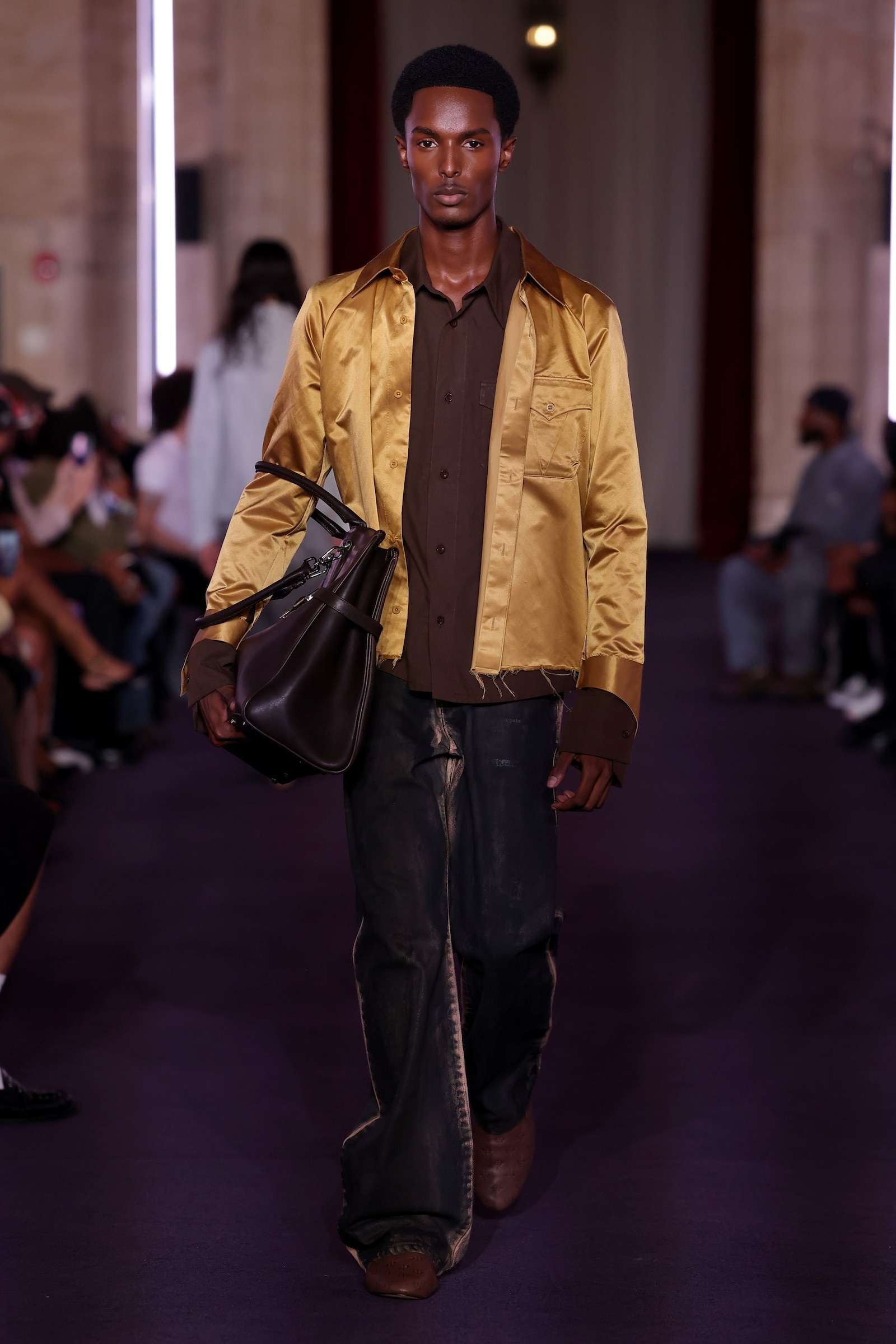 A Black male model in a gold satin jacket carries an oversized dark leather bag on the VIVET runway