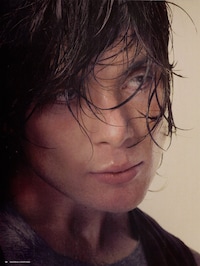 A portrait photograph of a person with dark, tousled hair and glasses, shot against a warm beige backdrop.